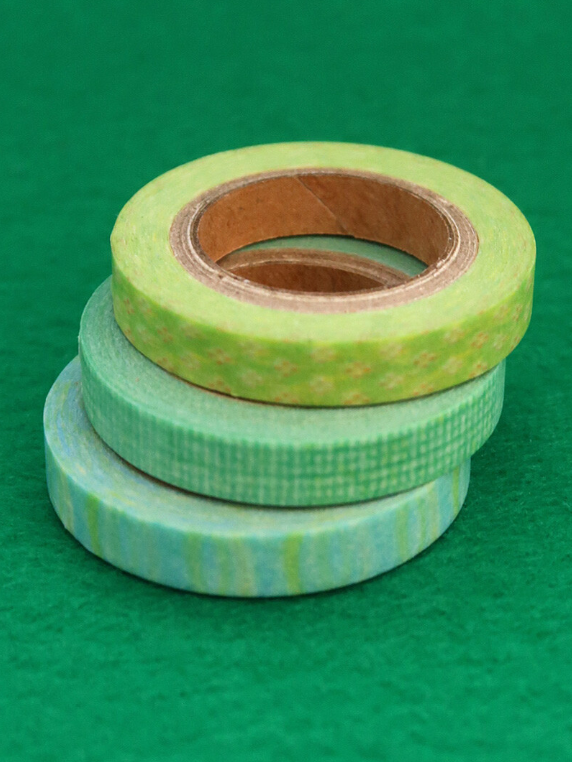 Three rolls of decorative washi tape in shades of green and light blue, stacked on a solid green background.