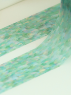 A close-up of a roll of washi tape with a textured, painterly pattern in shades of green, blue, and purple, unrolled diagonally across a white surface.
