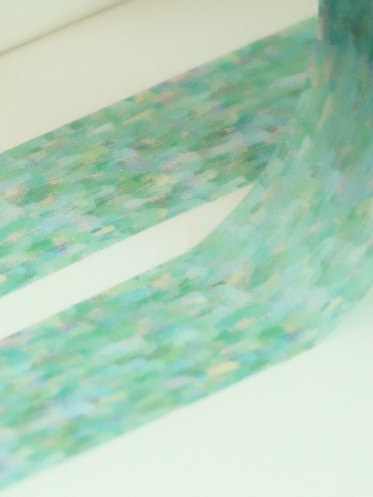 A close-up of a roll of washi tape with a textured, painterly pattern in shades of green, blue, and purple, unrolled diagonally across a white surface.