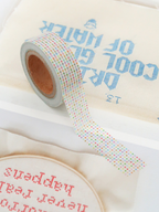 A close-up of a roll of washi tape with a pastel rainbow dotted pattern, laid diagonally over cream-colored embroidered fabric pieces with blue and red stitched text.