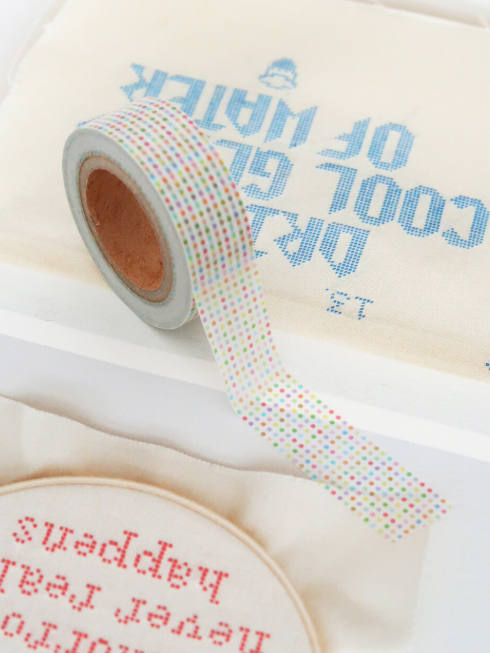 A close-up of a roll of washi tape with a pastel rainbow dotted pattern, laid diagonally over cream-colored embroidered fabric pieces with blue and red stitched text.
