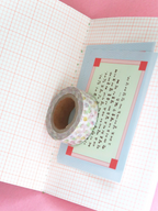 A roll of floral-patterned washi tape rests on a light blue card with Japanese calligraphy, placed on an open notebook with pink grid paper against a pink background.