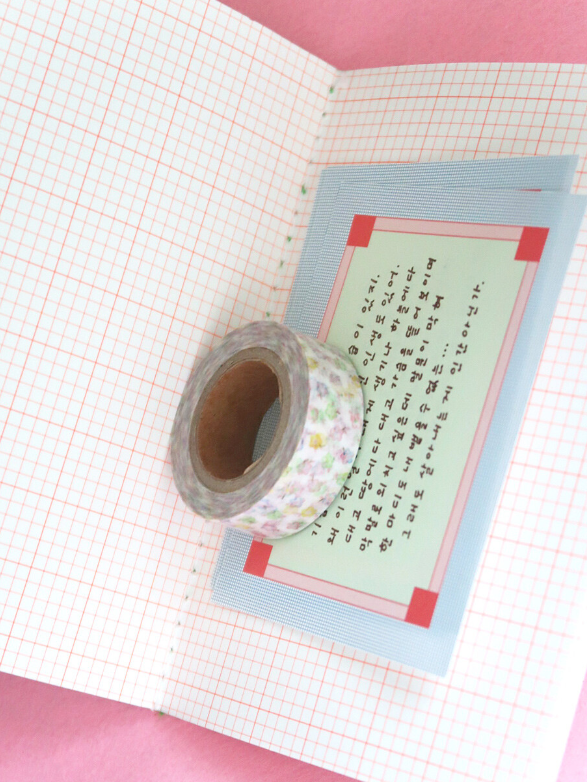 A roll of floral-patterned washi tape rests on a light blue card with Japanese calligraphy, placed on an open notebook with pink grid paper against a pink background.