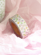 A roll of washi tape with a pastel multicolor floral pattern on a white background, resting on pink tissue paper.