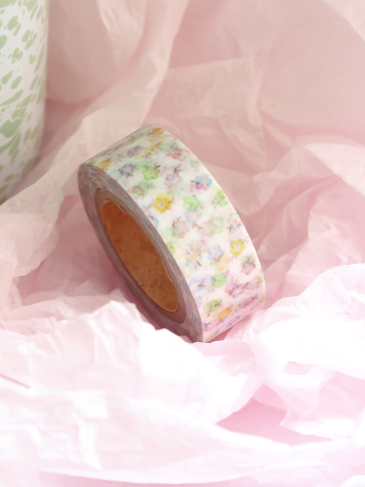 A roll of washi tape with a pastel multicolor floral pattern on a white background, resting on pink tissue paper.