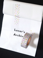 A roll of washi tape with a pastel multicolor floral pattern is shown partially unrolled on a white sheet of paper against a black background. The text 'Loner's Archiv' is printed on the sheet, and the tape roll itself is positioned in the lower right.