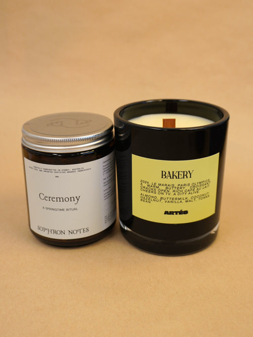 Two artisan candles from the brand Artés on a warm beige background. On the left is the 'Ceremony' loose leaf tea in a dark glass jar with a silver lid and white label.