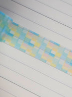 A close-up of a pastel checkered patterned washi tape, featuring light blue, yellow, and pink squares, laid diagonally across a white lined notebook.