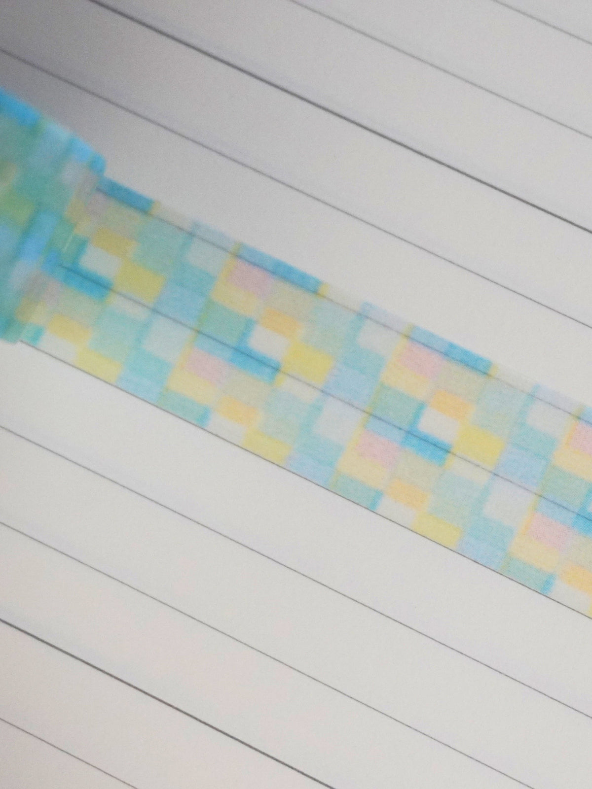 A close-up of a pastel checkered patterned washi tape, featuring light blue, yellow, and pink squares, laid diagonally across a white lined notebook.